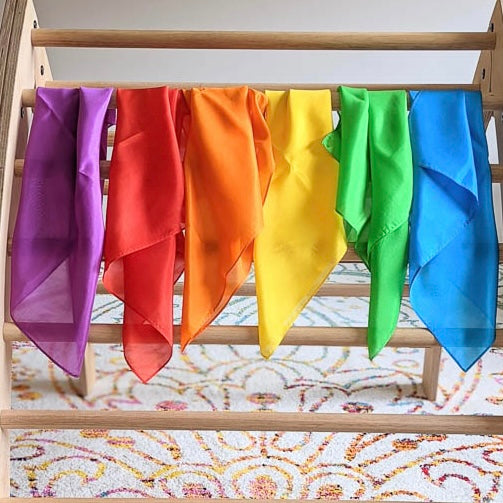 Colorful mini silk scarves hanging on a wooden pickler against a patterned rug.