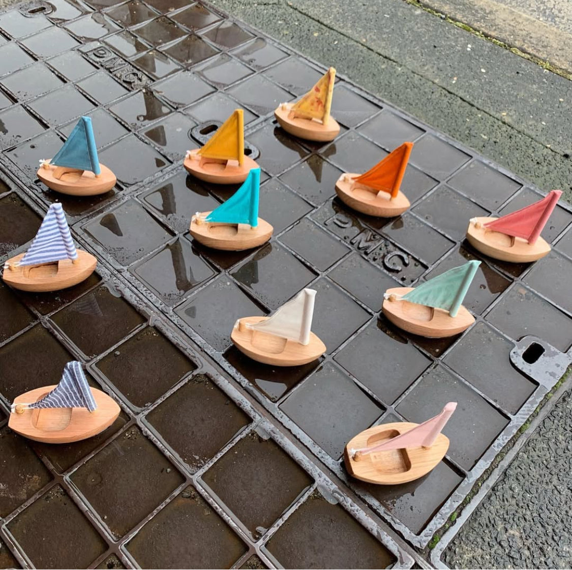 a variety of newly arrived toy wooden sailboats on the pavement at the front of Lyreird