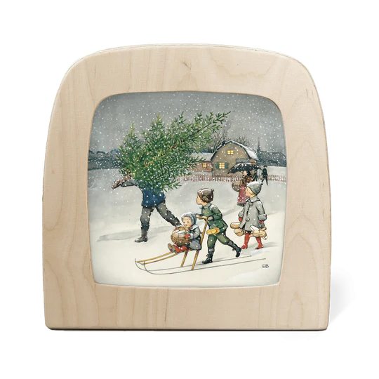 A wooden silhouette screen featuring an illustration of a European Winter scene with a man carrying a firtree home in the snow with three children alongside him