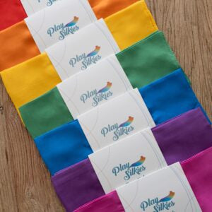 Colourful rainbow of shades of silk fabric play cloths with 'Play Silky' branding on white card wrapping, displayed on a wooden surface