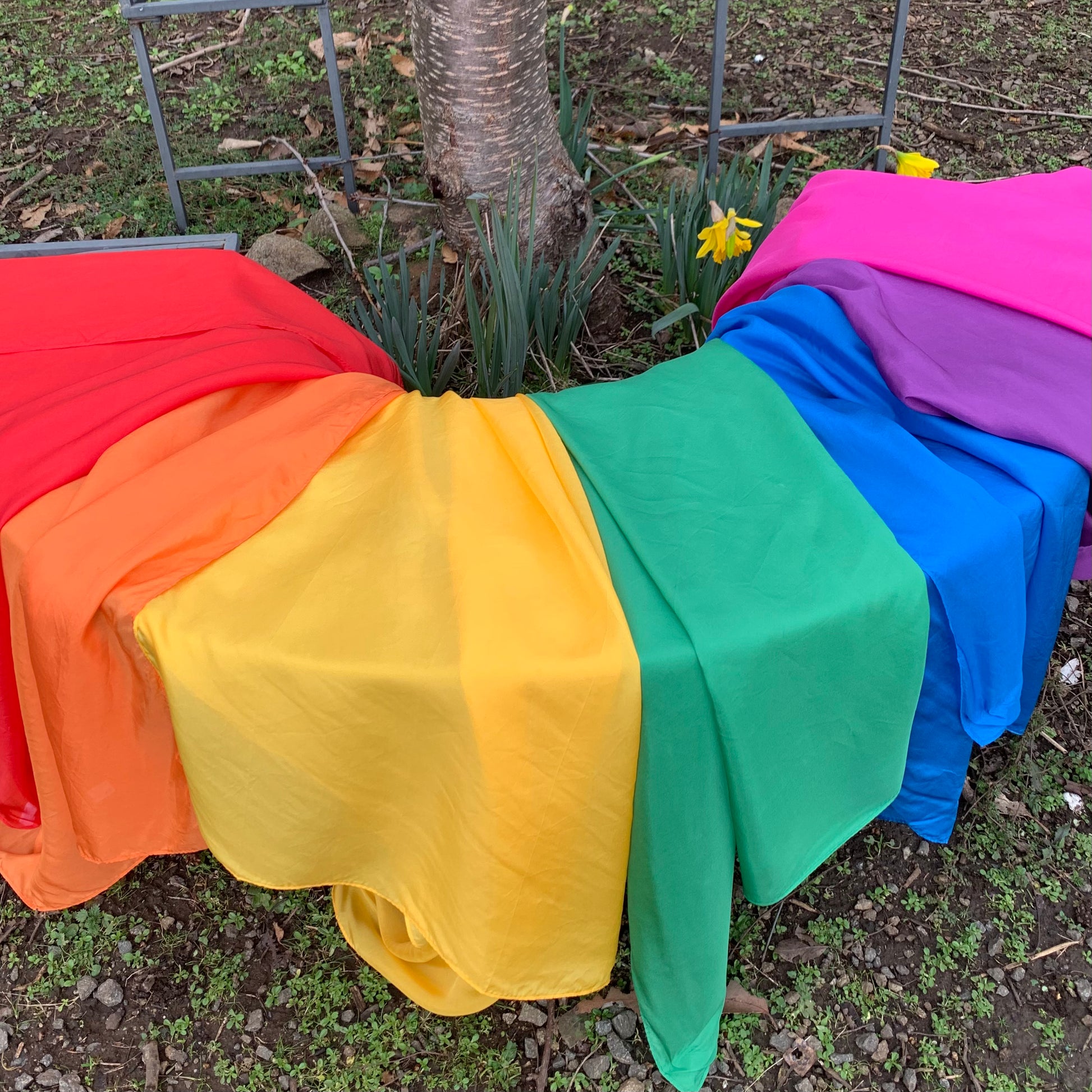 a set of colourful silk play cloths draped in rainbow order over an outdoor garden bench outdoors