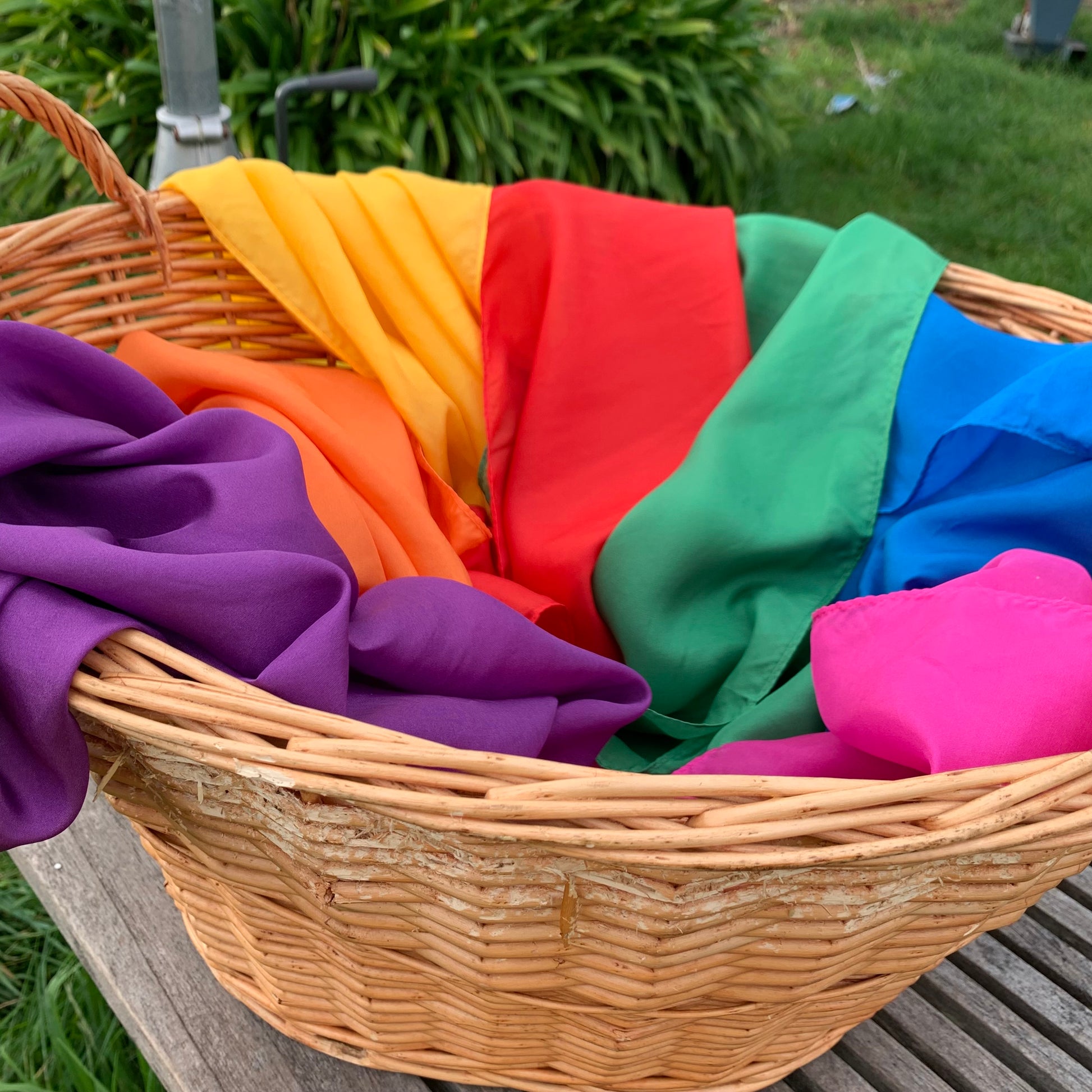 Wicker basket filled with colourful silk cloths in a rainbow of colour, sitting on a wooden table under the clothesline.