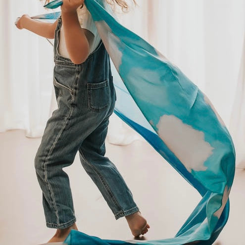 child denim overalls running  whilst holding a large blue silk fabric in a minimal indoor setting