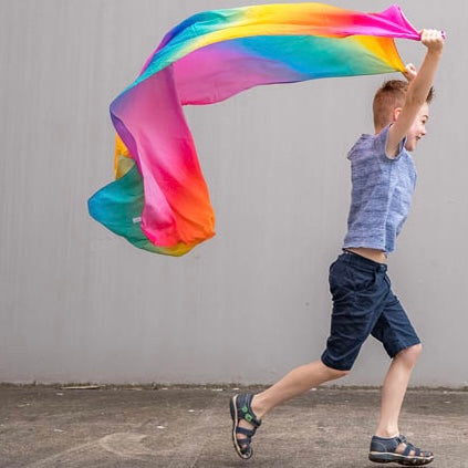 boy running whilst holding a bright colorful silk rainbow scarf against a plain background