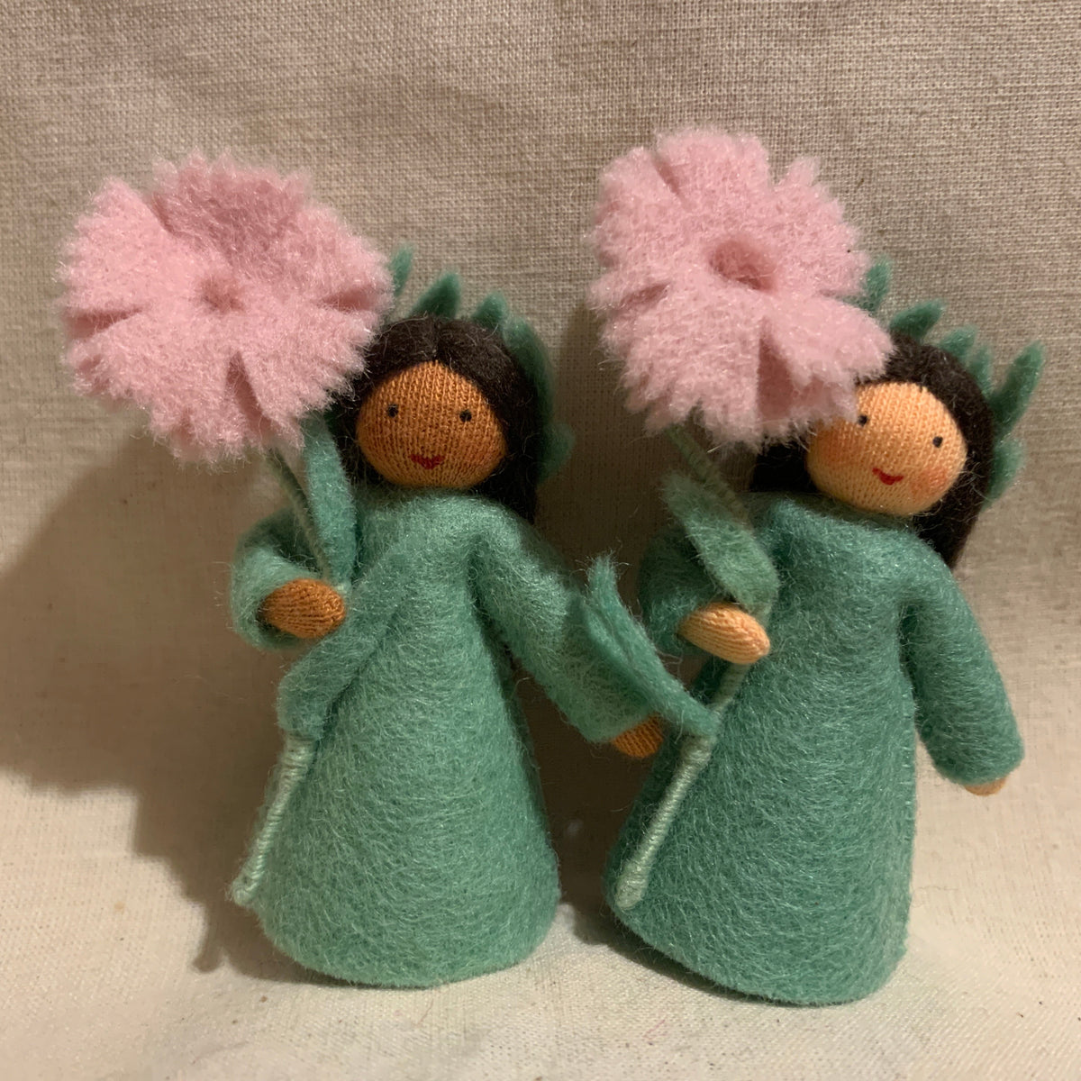 Two handmade felt flower dolls with pink carnation petals, green felted dresses, and dark brown felted hair, standing side by side against a neutral beige fabric backdrop.