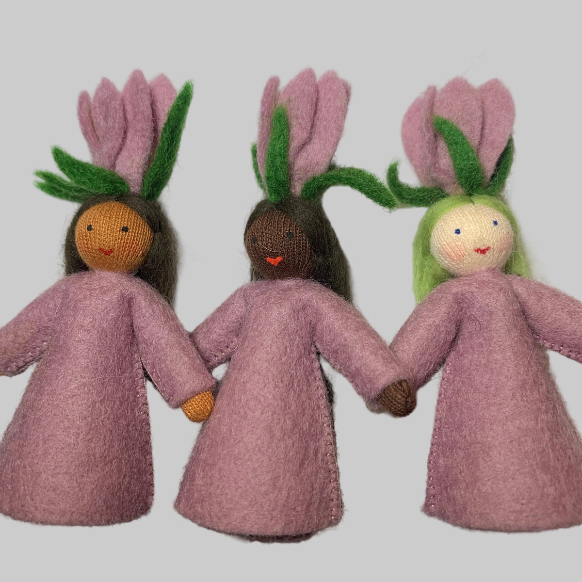 Three handmade felt dolls wearing pink dresses and flower-like hair ornaments, arranged side by side on a white sheet of paper.