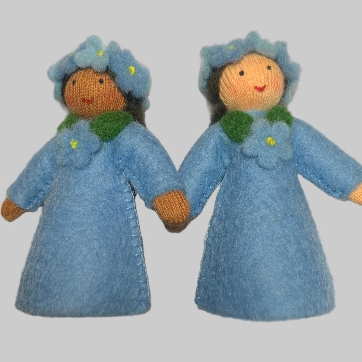 Two small handmade forgetmenot fairy felt dolls wearing matching light blue dresses and hats, standing side by side on a white surface.
