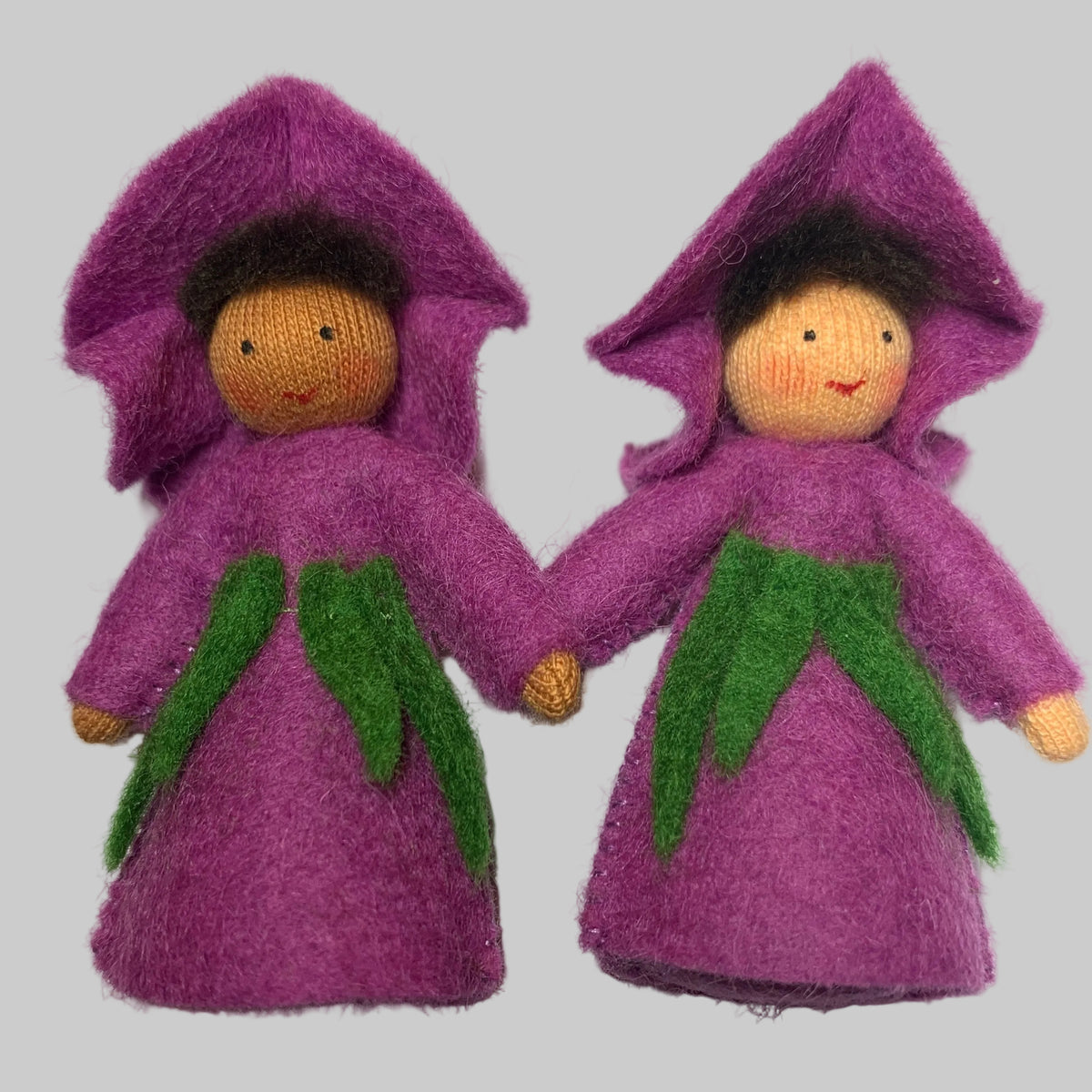 Two small felted wool fairy dolls wearing purple pointed hats and dresses with green leaf accents, standing side by side on a white surface.
