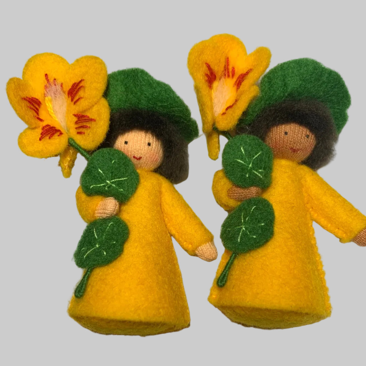 Two handmade felt garden nasturtium fairy dolls dressed in yellow cloaks and green hats, each holding a yellow flower with red and orange details, displayed on a white surface.