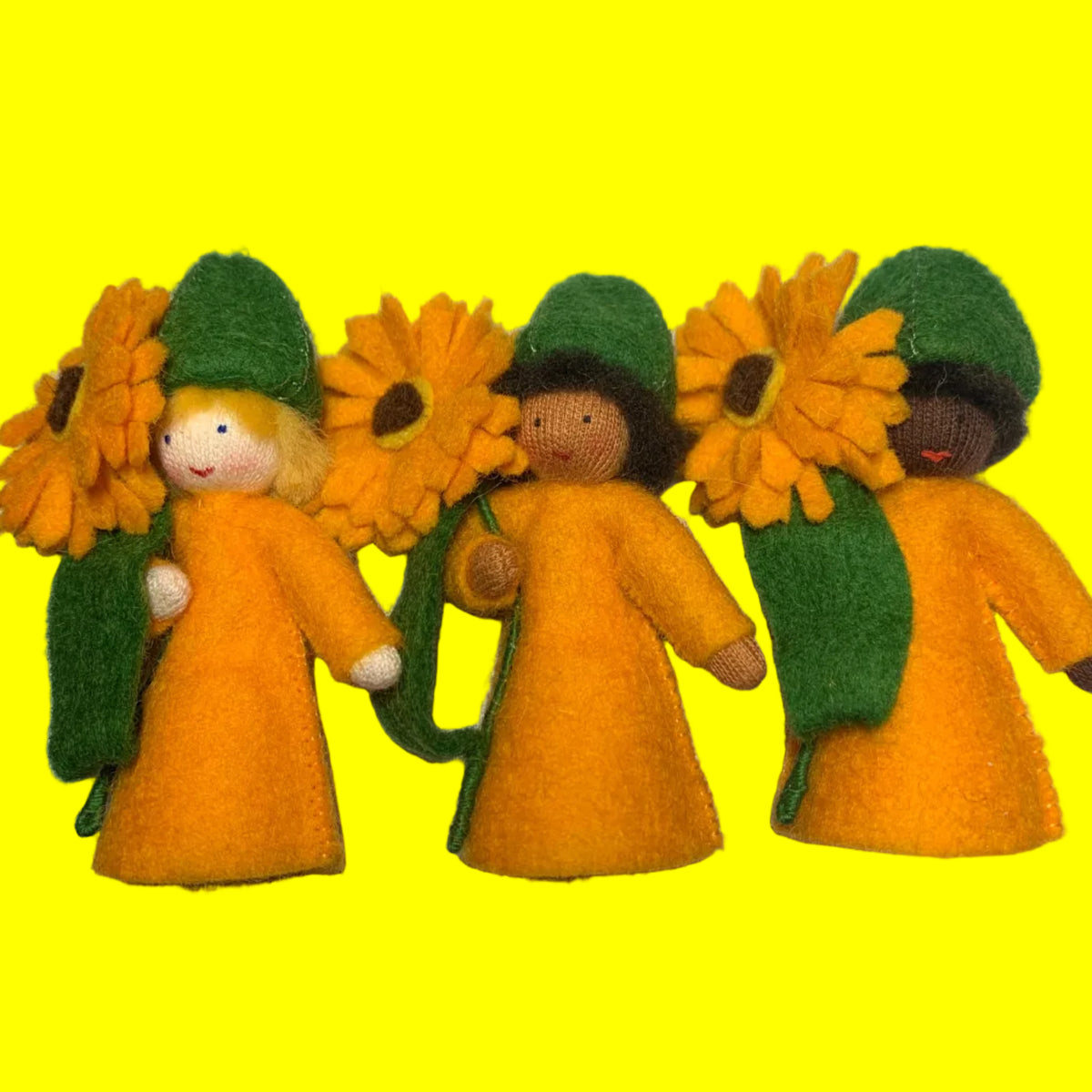 Three small handmade calendula felt dolls wearing green hats and long yellow-orange dresses, each holding a large yellow sunflower, arranged on a white surface.