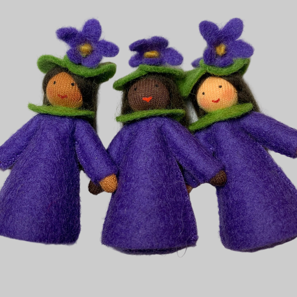 Three small felt dolls wearing purple dresses and flower hats, arranged side by side on a plain light background.