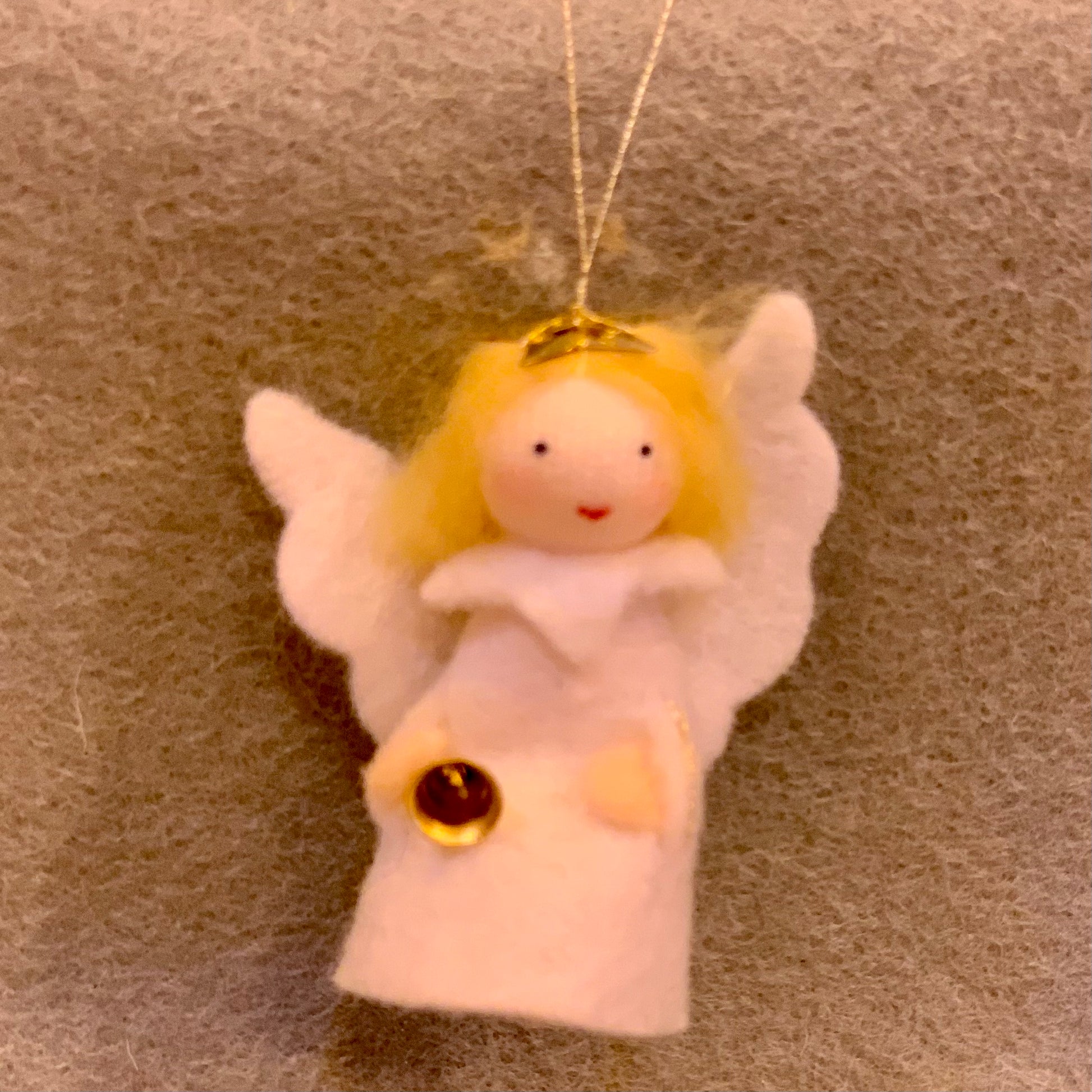 Angel ornament with blonde hair and white wings holdingh a tinkling bell on a grey background