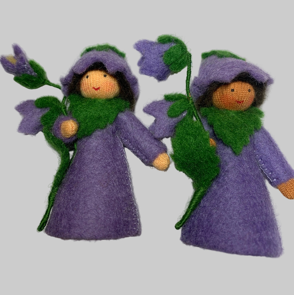 Two small felted wool fairy dolls dressed in purple cloaks with green leafy collars and purple flower motifs, displayed on a white surface.