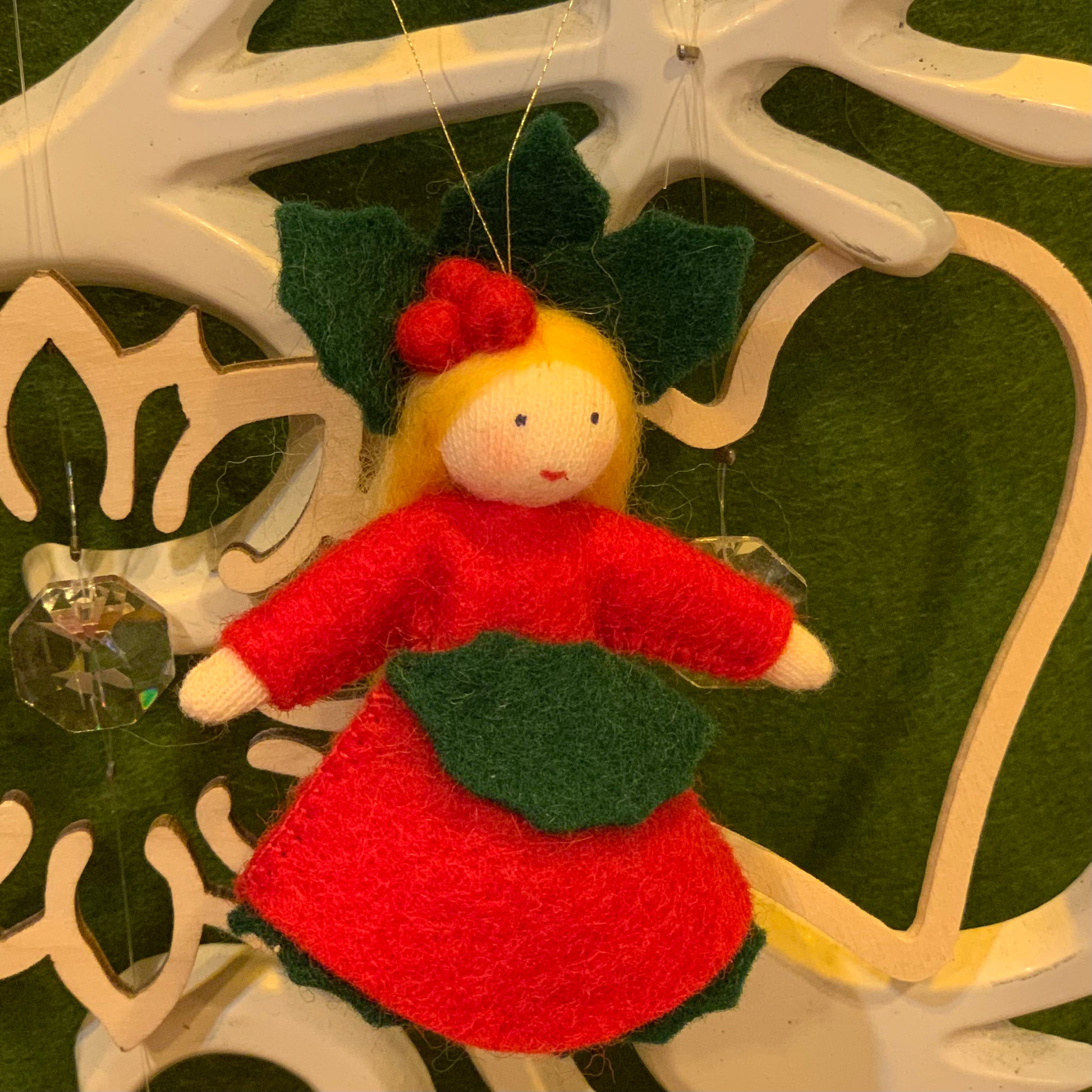 Felt Christmas ornament of a holly fairy in a red dress with green leaves, hanging on a decorative white stand.