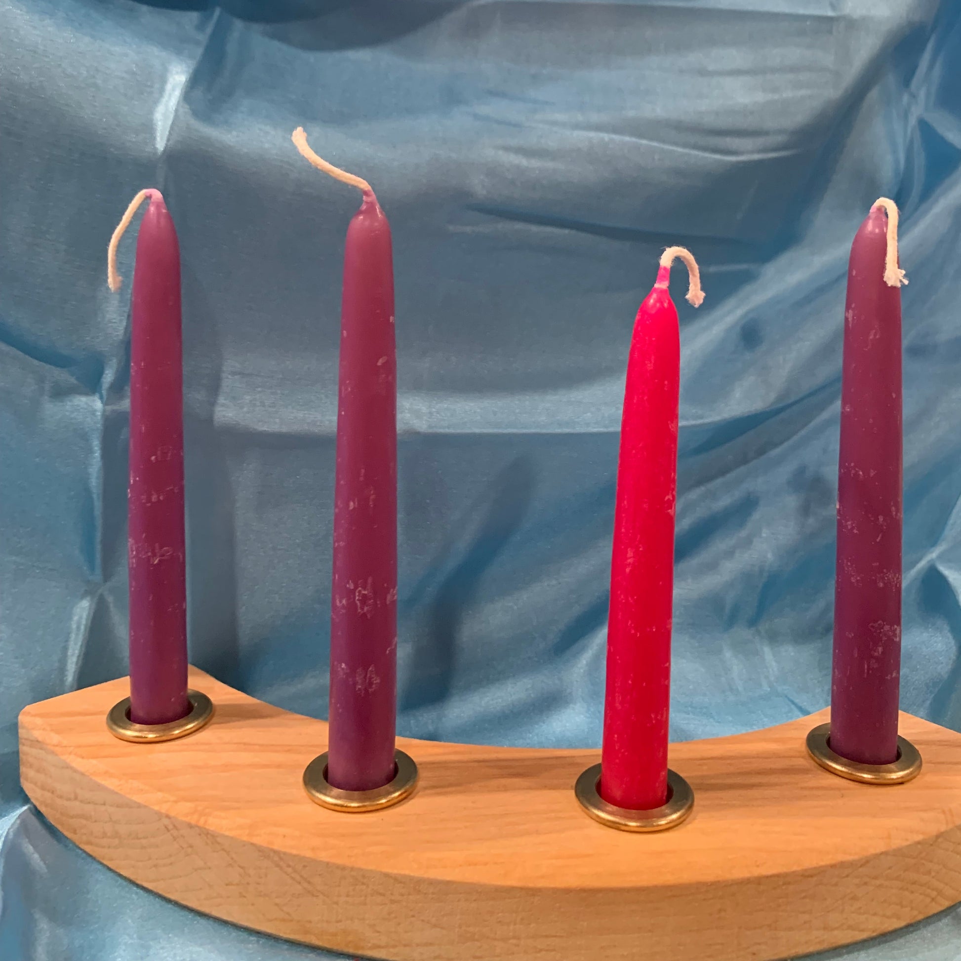 A set of four artisan beeswax candles , catholic tradition of x3 purple and one pink , arranged in a wooden holder, in brass cupsagainst a blue silk background.
