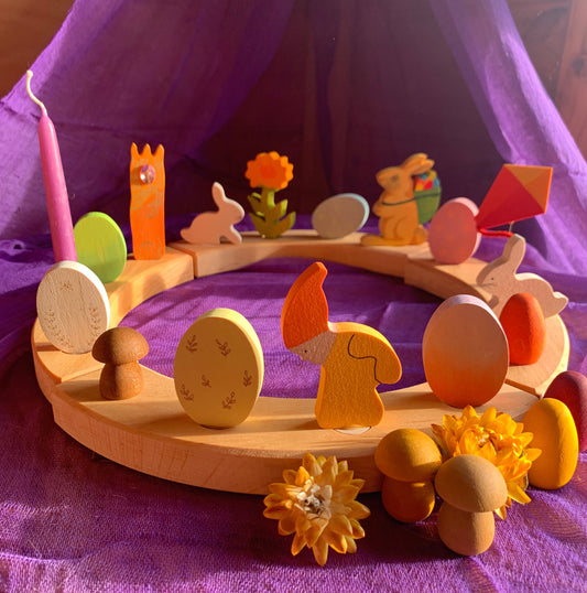 A wooden celebration ring decorated with colorful Easter-themed wooden figures, including bunnies, eggs, and other Easter -related shapes, displayed on a purple background.