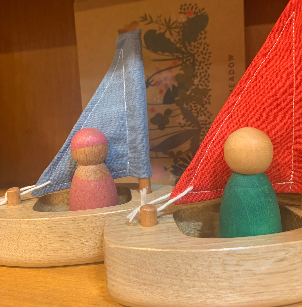 A wooden toy boat with fabric sails in red, blue, featuring wooden peg people sailors.