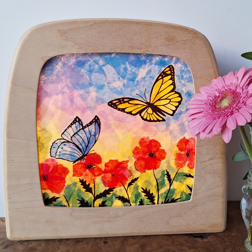 Wooden picture frame with a colourful tissue paper decoupage  of butterflies and flowers, next to a vase with flowers.