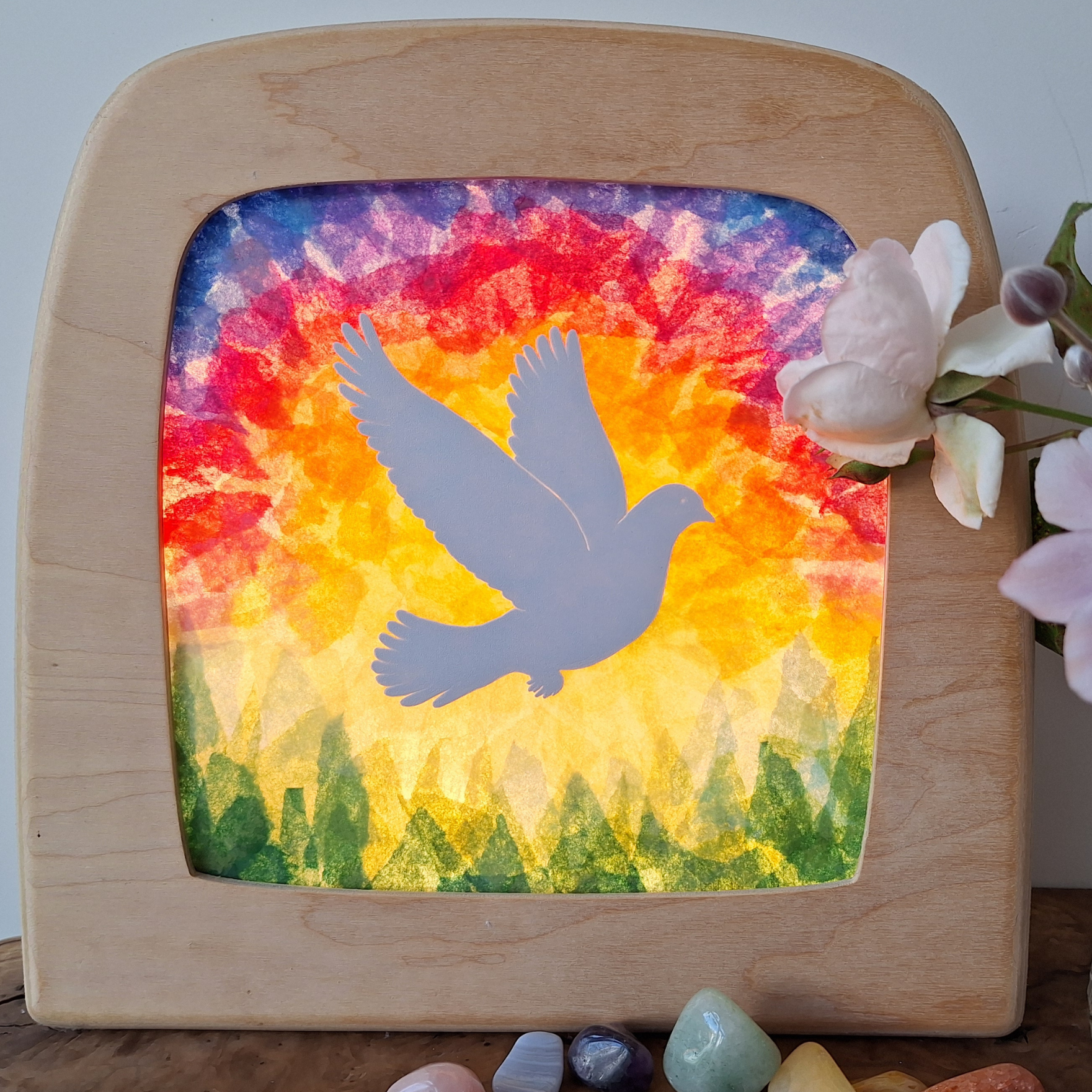 Colorful tissue paper art design with a bird silhouette in a wooden frame, surrounded by flowers and stones.