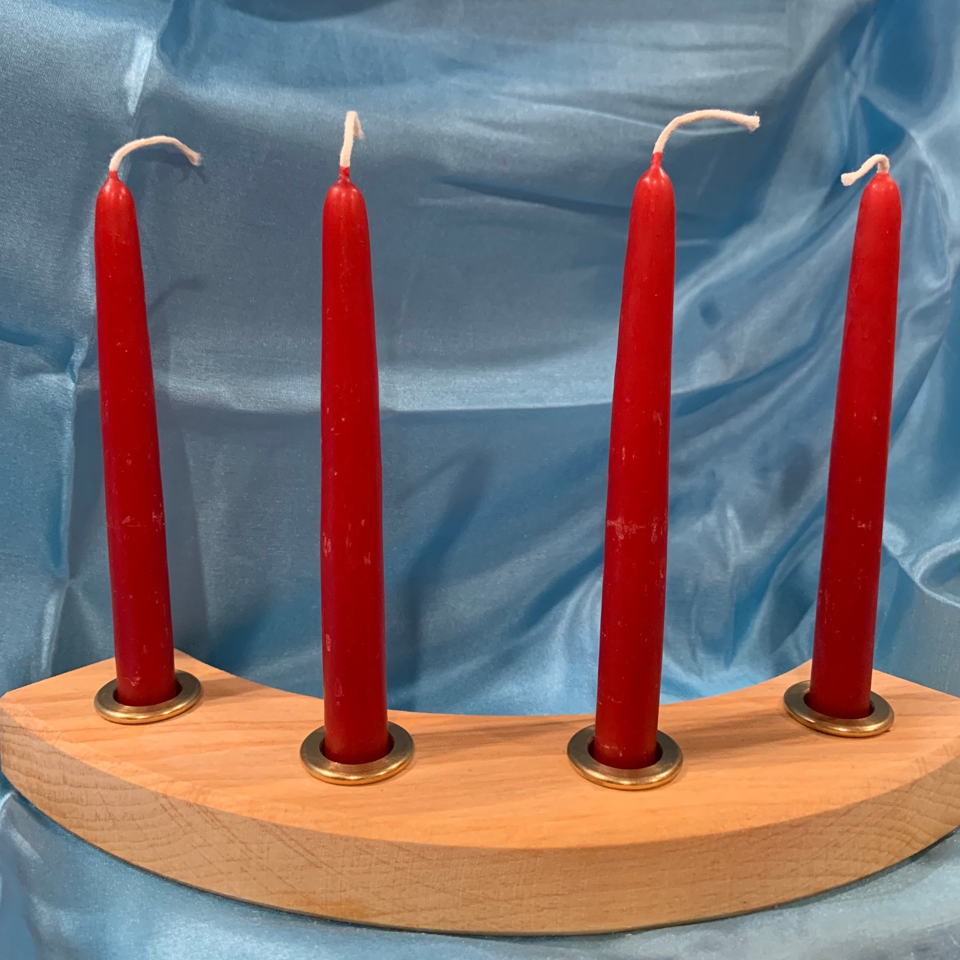 A set of four red artisan beeswax candles arranged in brass cups in a wooden holder, , displayed against a blue silk background.